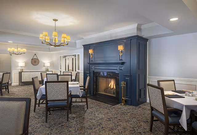 A cozy dining room with several tables covered in white tablecloths and surrounded by upholstered chairs. A dark blue fireplace with a lit fire is the focal point of the room, flanked by two wall sconces. The room features patterned carpet, soft lighting from chandeliers, and neutral-colored walls with white wainscoting.