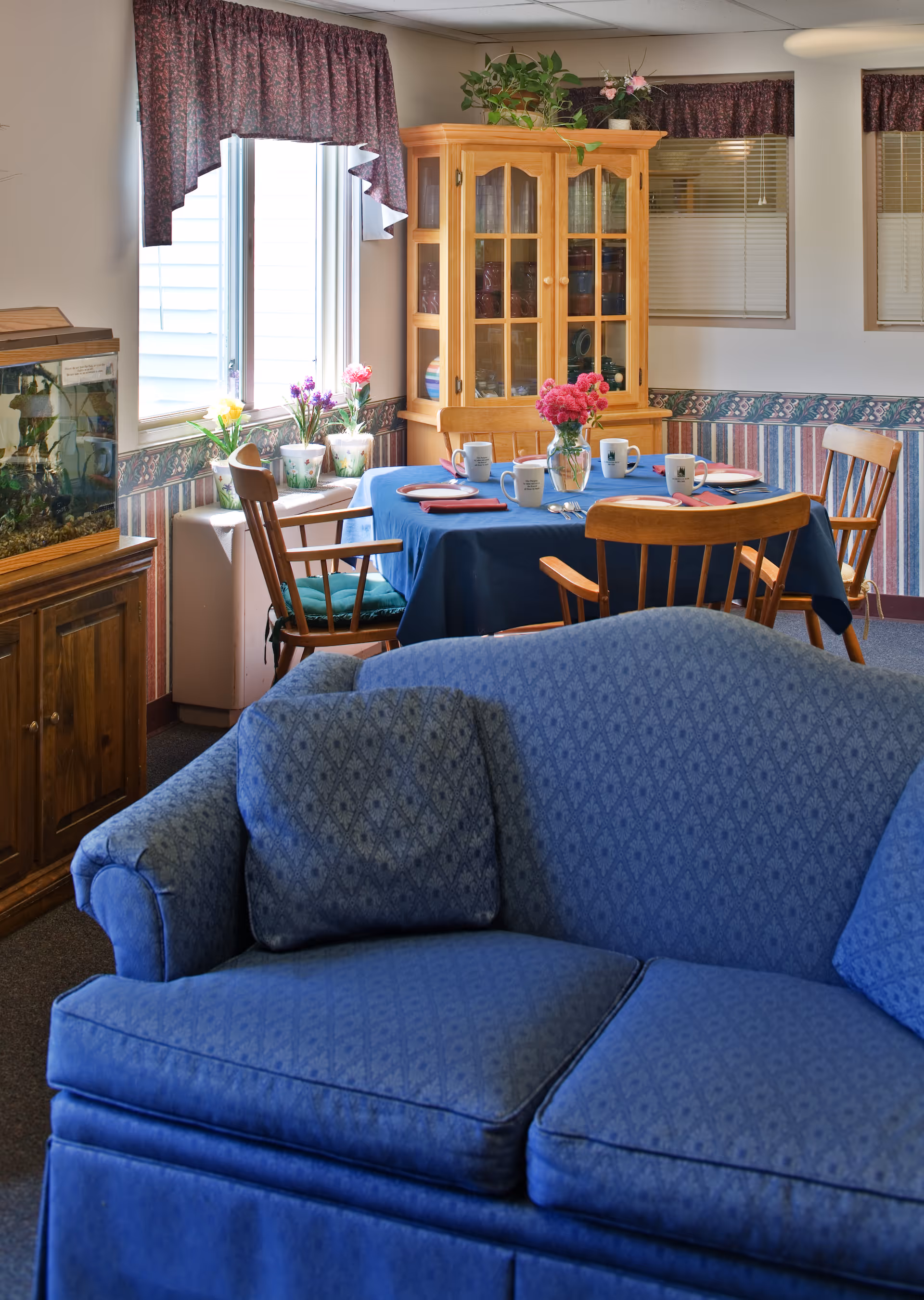 Cozy senior living common room featuring a blue sofa in the foreground and a dining table set with wooden chairs and a glass-front cabinet by the window.