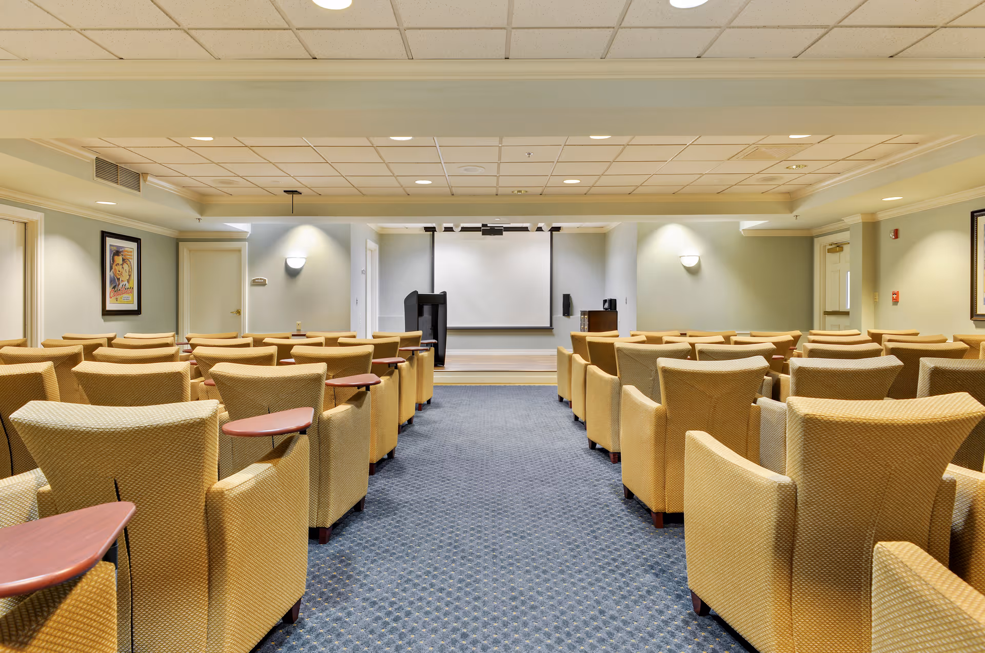 A conference or presentation room with rows of beige upholstered chairs featuring attached wooden writing surfaces, facing a small stage with a podium and a large white projection screen. The room has a blue carpet with a subtle pattern, light-colored walls, and framed artwork on the side walls.