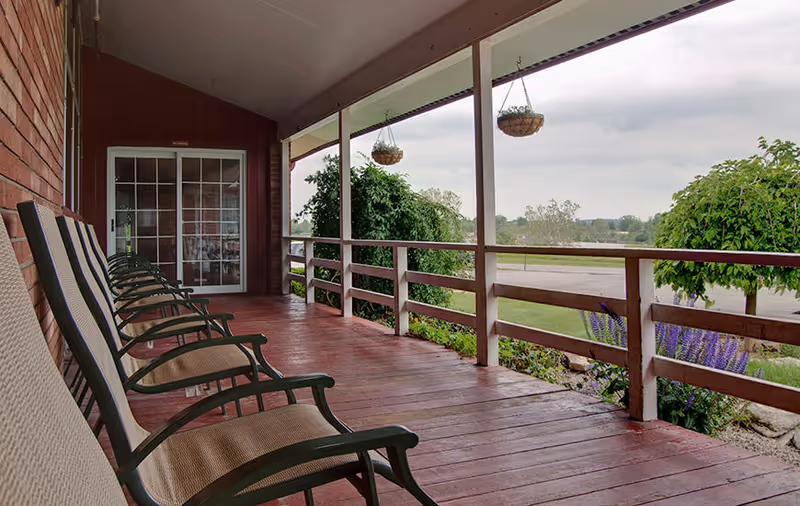 A covered wooden porch with several cushioned chairs lined up facing outward. The porch has hanging flower baskets and overlooks a garden area with bushes and purple flowers. The sky is cloudy and there is a glass door leading inside the building.
