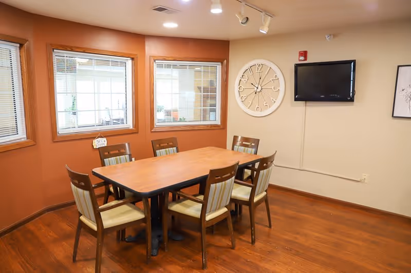 A small dining room with a rectangular wooden table surrounded by six chairs with striped cushions. The room has three windows with blinds, an analog wall clock, a mounted flat-screen TV, and a framed picture on the wall. The floor is wooden and the walls are painted in two tones, orange and beige.