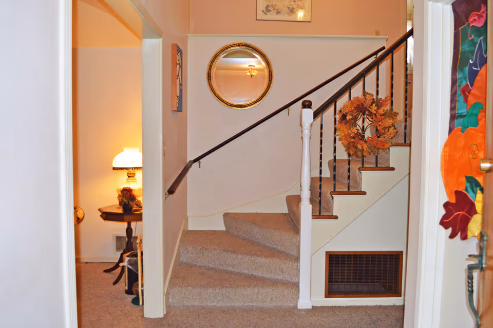 Interior view of a carpeted staircase with a wooden handrail and black balusters. A decorative autumn wreath is hanging on the staircase railing. To the left, there is a small room with a lit table lamp on a wooden side table. A round mirror with a gold frame is mounted on the wall above the staircase. The walls are painted in light colors, and there is a colorful fall-themed decoration hanging on the right side near the door.