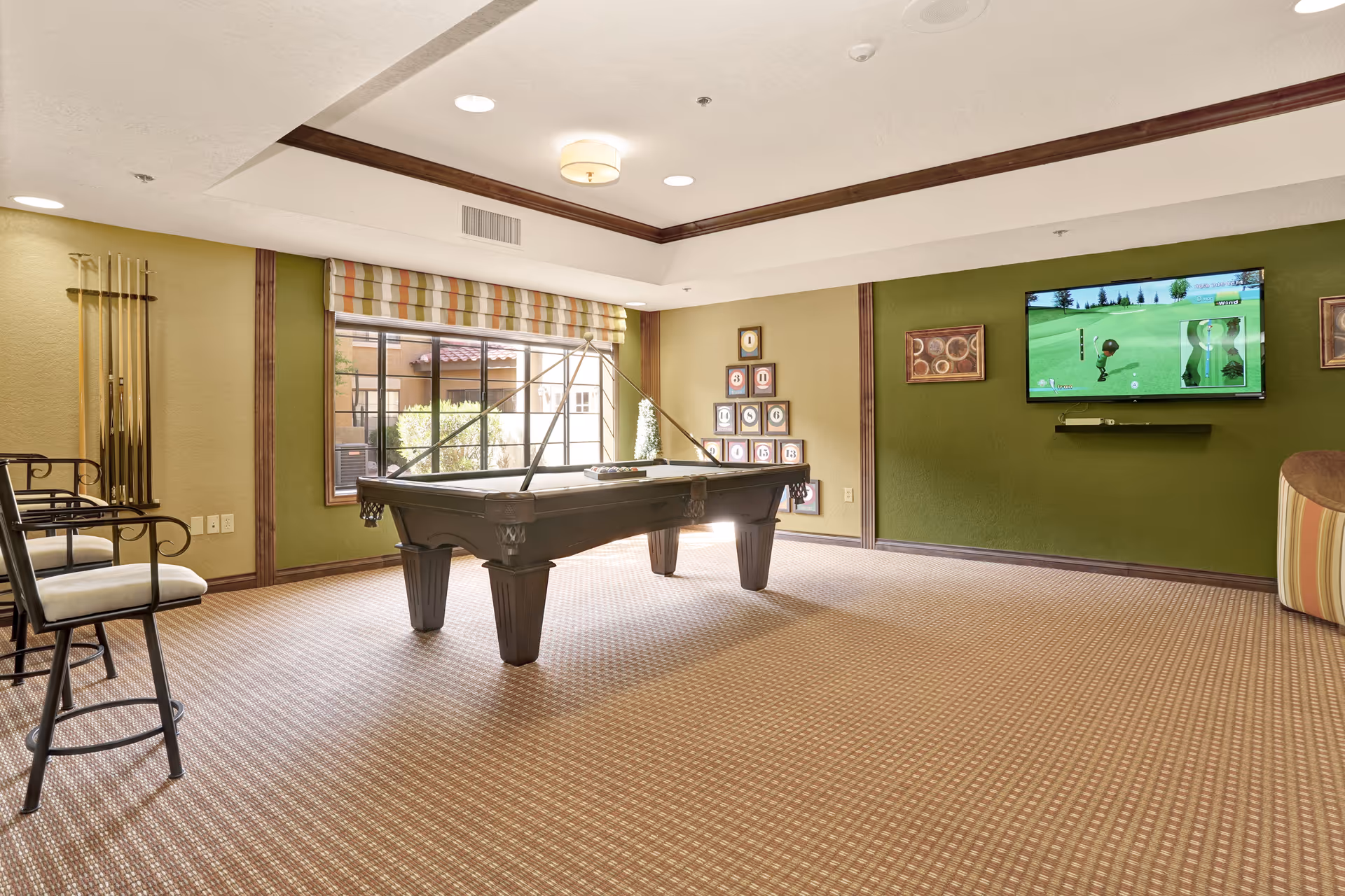 A bright recreation room with a pool table centered, cue racks, seating, and a TV mounted on a green wall.