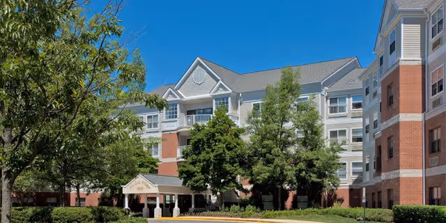 Exterior view of a multi-story senior living facility named Pin Oak Village, featuring a combination of brick and light-colored siding, surrounded by green trees and landscaping under a clear blue sky.