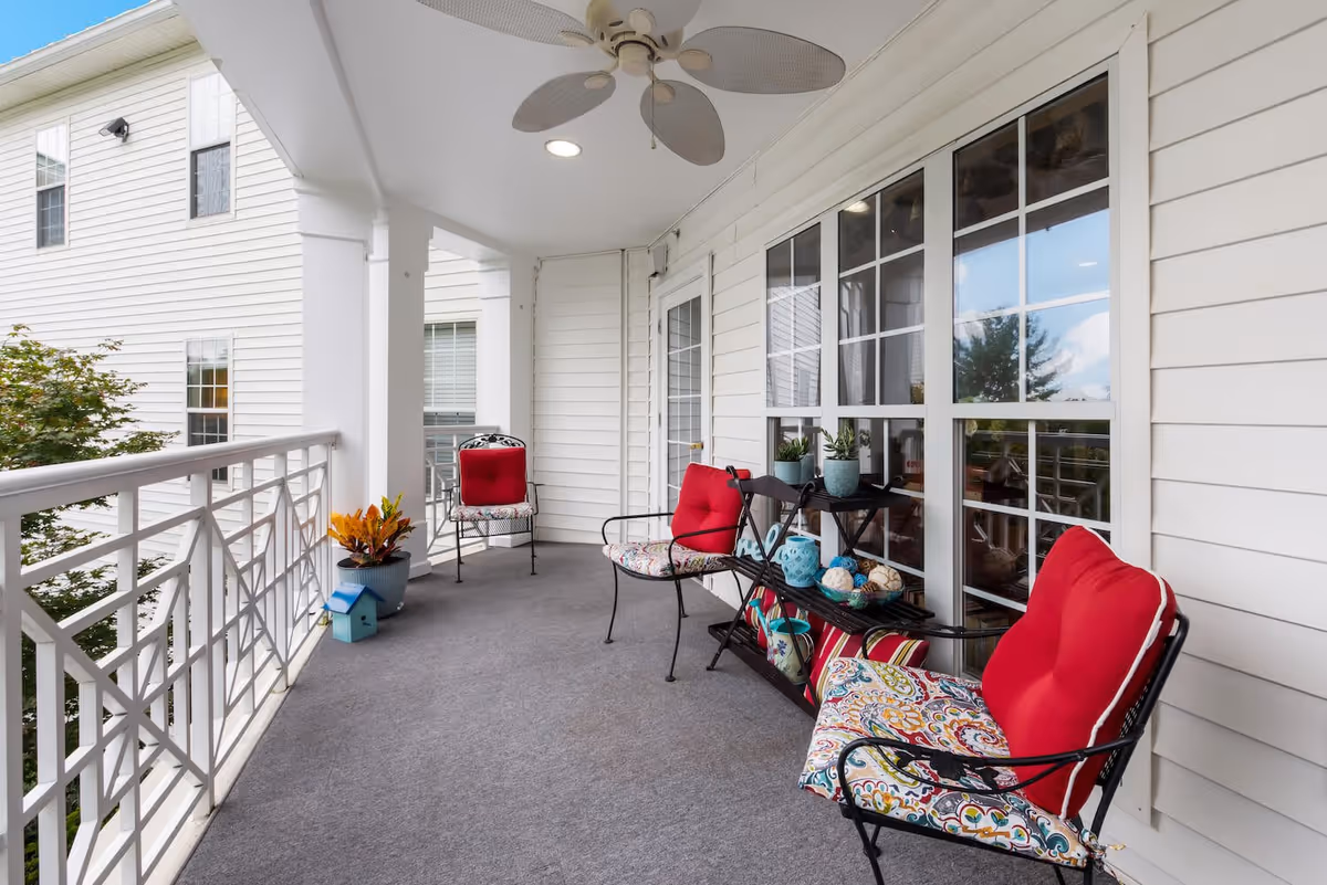 A covered outdoor balcony area with white railings and walls, featuring three metal chairs with colorful cushions, a small black shelf with decorative items and plants, a ceiling fan, and a view of trees and neighboring building windows.