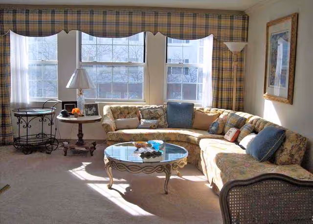 A bright living room with large windows covered by plaid valances and curtains. There is a curved floral-patterned sofa with multiple cushions, a glass coffee table with decorative items, a side table with a lamp and framed photos, and a metal cart near the window. A framed picture hangs on the wall.