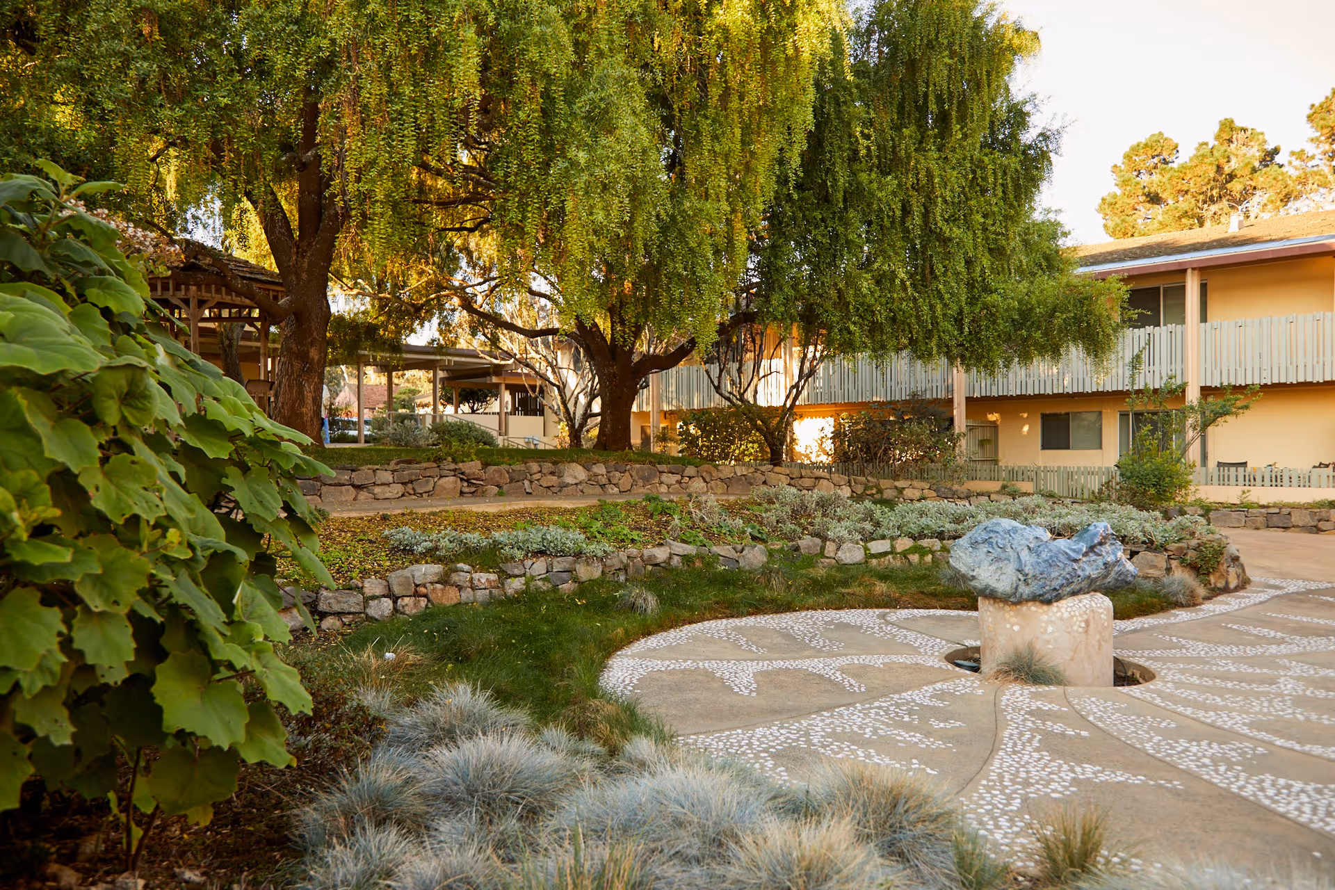 Courtyard garden with a circular patterned patio and stone sculpture, surrounded by trees, landscaping, and a two-story residential building.
