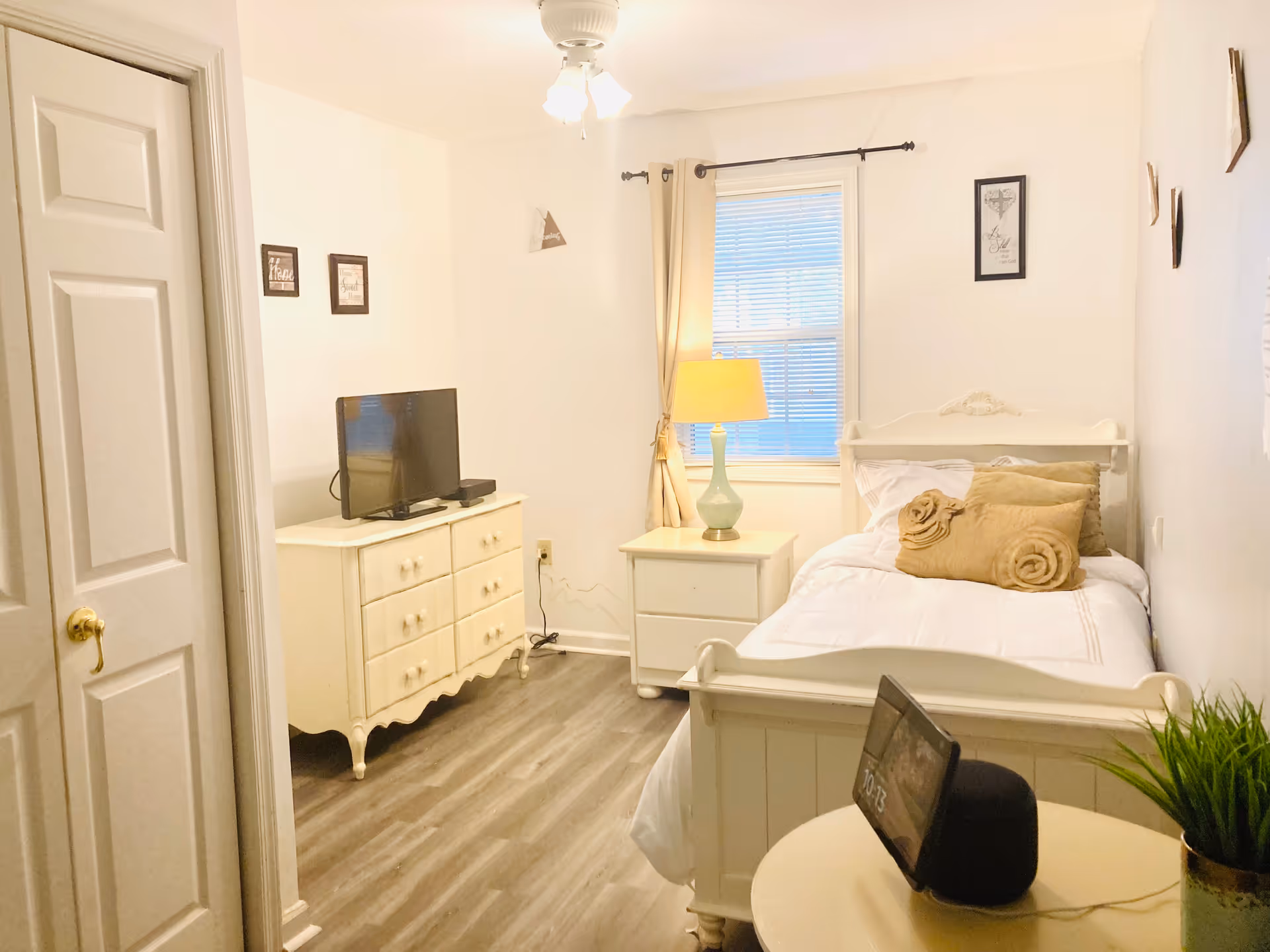 A cozy bedroom with a single white bed adorned with beige pillows and a rolled towel. Next to the bed is a white nightstand with a green lamp. Across from the bed is a white dresser with a flat-screen TV on top. The room has light-colored walls, wood flooring, a window with beige curtains, and a ceiling fan. A small round table with a digital clock and a potted plant is in the foreground.