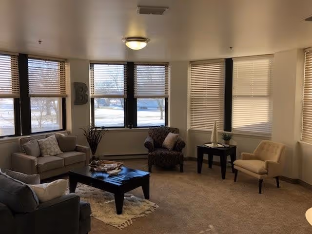 A cozy living room with multiple armchairs and a sofa arranged around a dark wooden coffee table on a beige carpet. Large windows with blinds allow natural light to fill the room. There is a small side table with decorative items and a letter 'B' mounted on the wall near the windows.