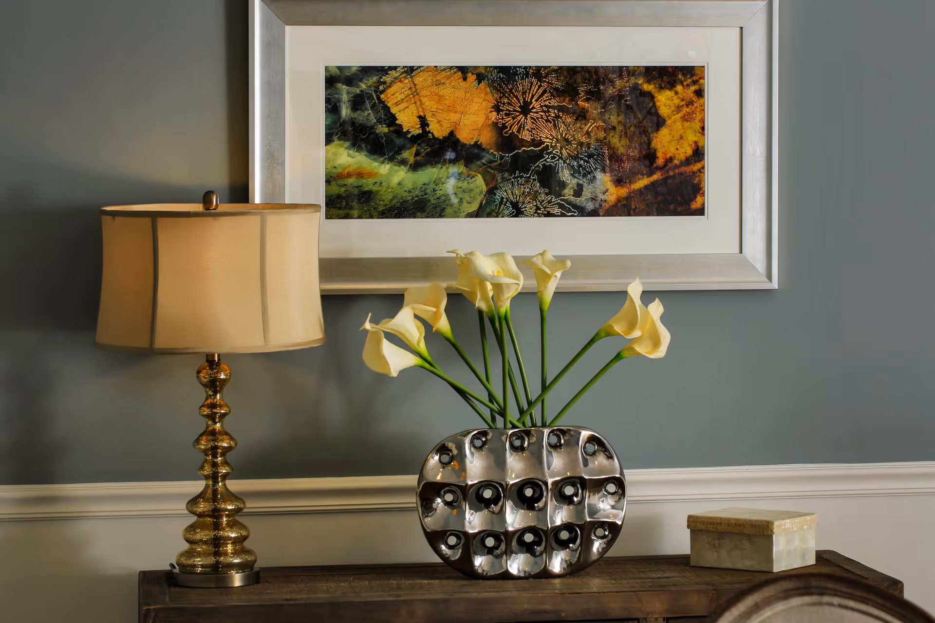A decorative setup on a wooden table featuring a gold-toned lamp with a beige lampshade, a modern silver vase holding white calla lilies, and a small rectangular box. Above the table hangs a framed abstract artwork with green, yellow, and orange hues on a gray wall with white wainscoting.