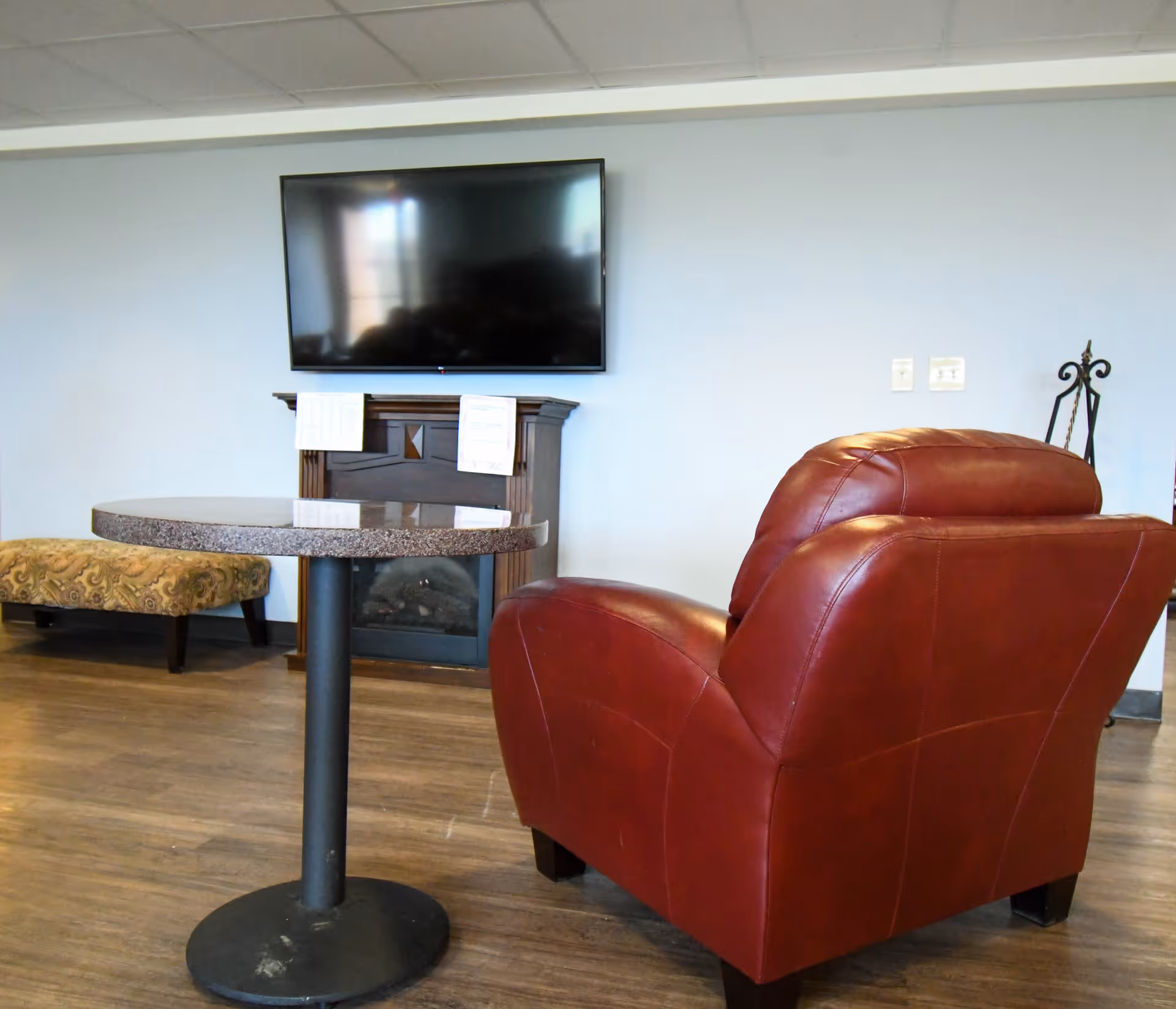 A cozy living room area with a red leather armchair, a round table with a black pedestal base, a patterned upholstered bench, a wall-mounted flat screen TV above a dark wooden electric fireplace, and a light blue wall background.