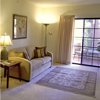 A cozy living room with a beige sofa adorned with multiple cushions, a wooden side table with a lamp and magazines, a floor lamp in the corner, a framed picture on the wall, a patterned area rug on the carpeted floor, and sliding glass doors leading to an outdoor patio with chairs.