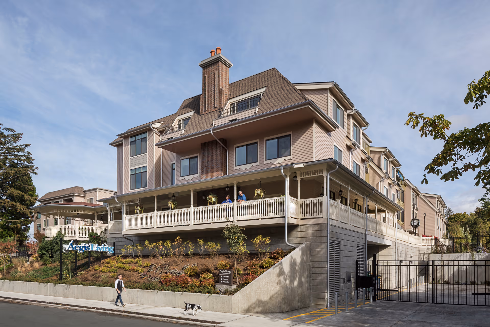 Multi-story senior living building with a wraparound porch, landscaped slope, and a person walking a dog on the sidewalk.