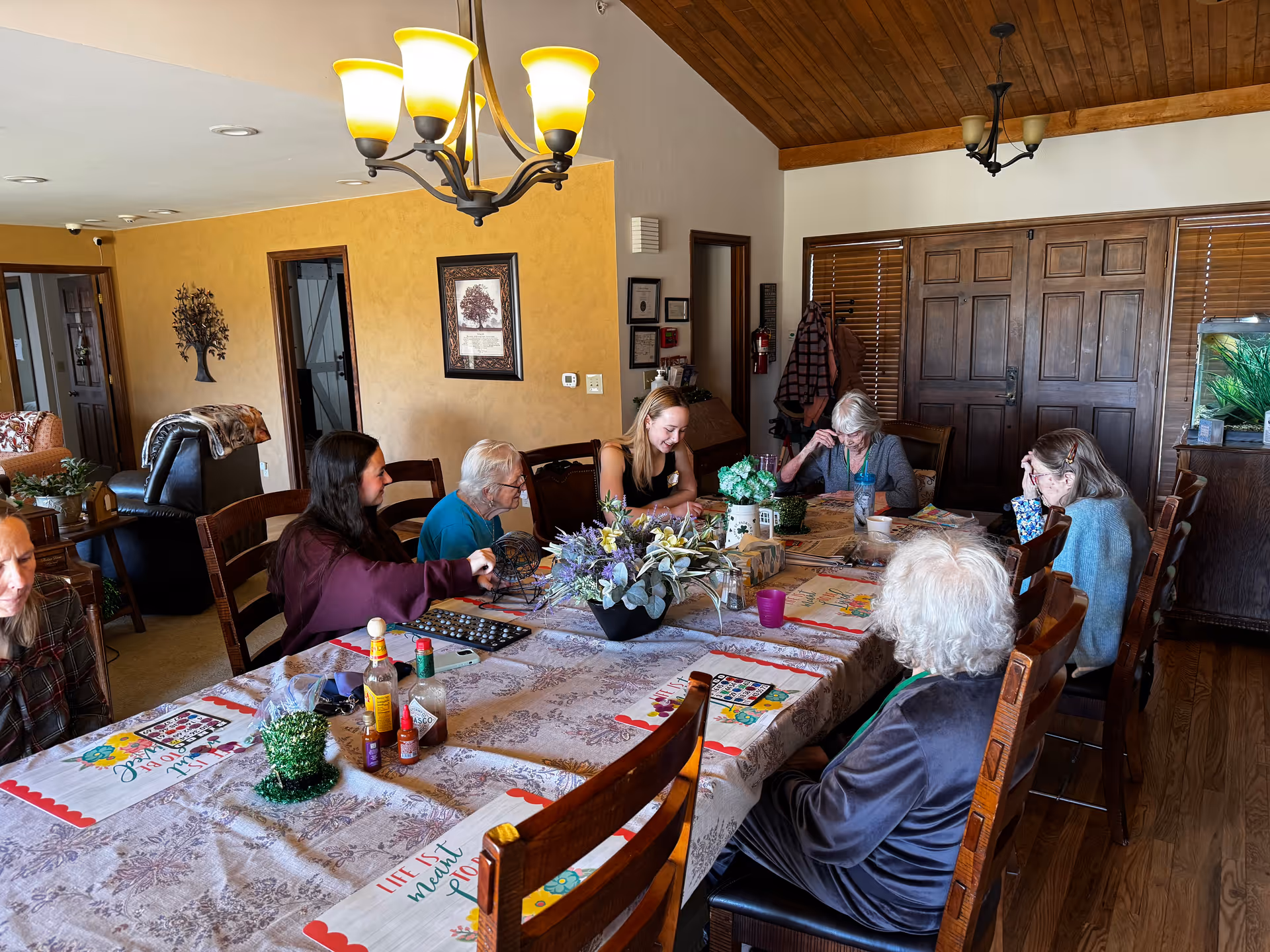 Several elderly residents and a caregiver sit around a long table playing games in a wood‑ceilinged communal dining room.