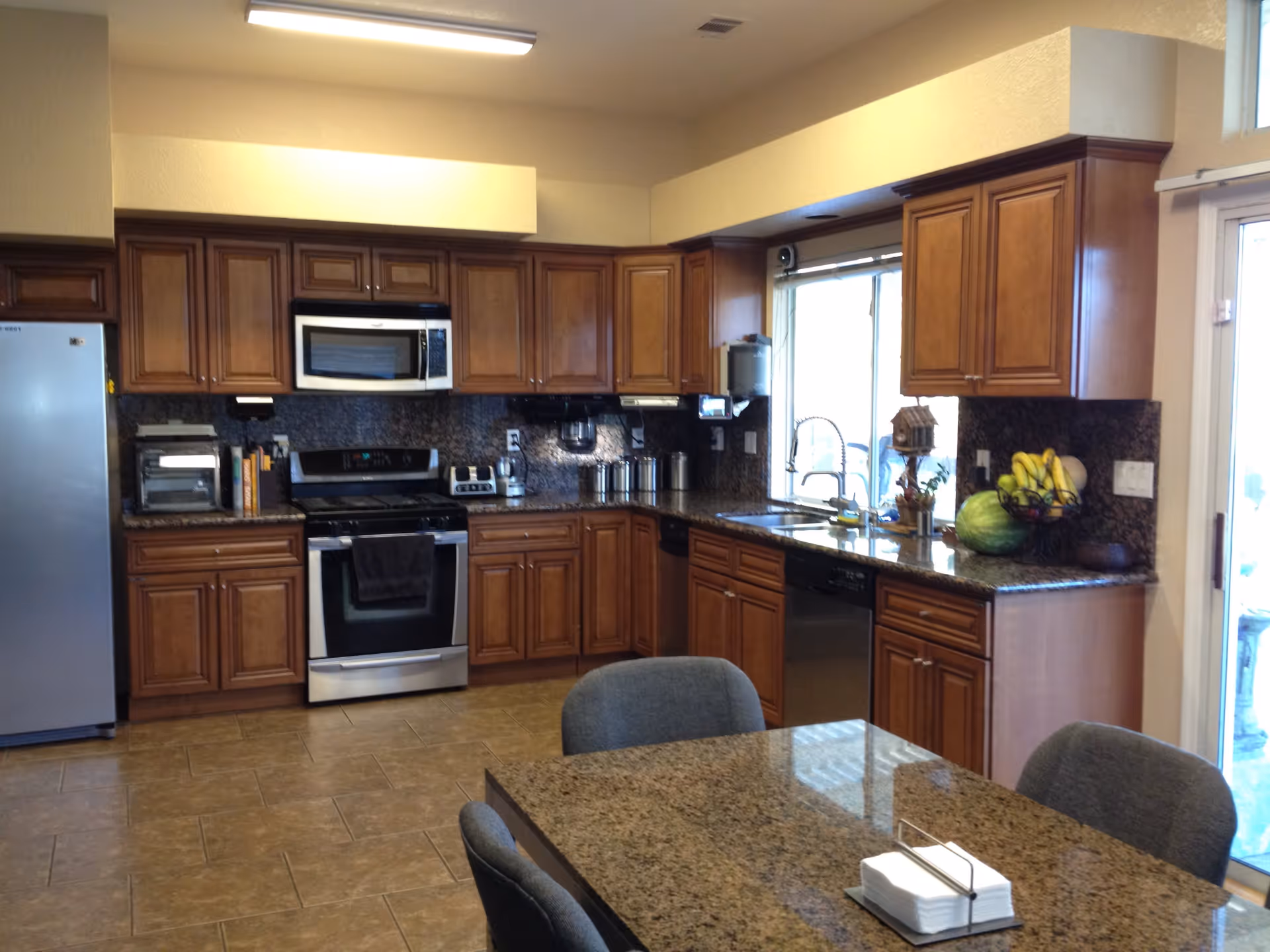 A modern kitchen with wooden cabinets, stainless steel appliances including a refrigerator, oven, microwave, toaster, and blender. There is a granite countertop with a sink under a window, and a dining table with gray chairs in the foreground. Various kitchen items and fruits are placed on the counter.
