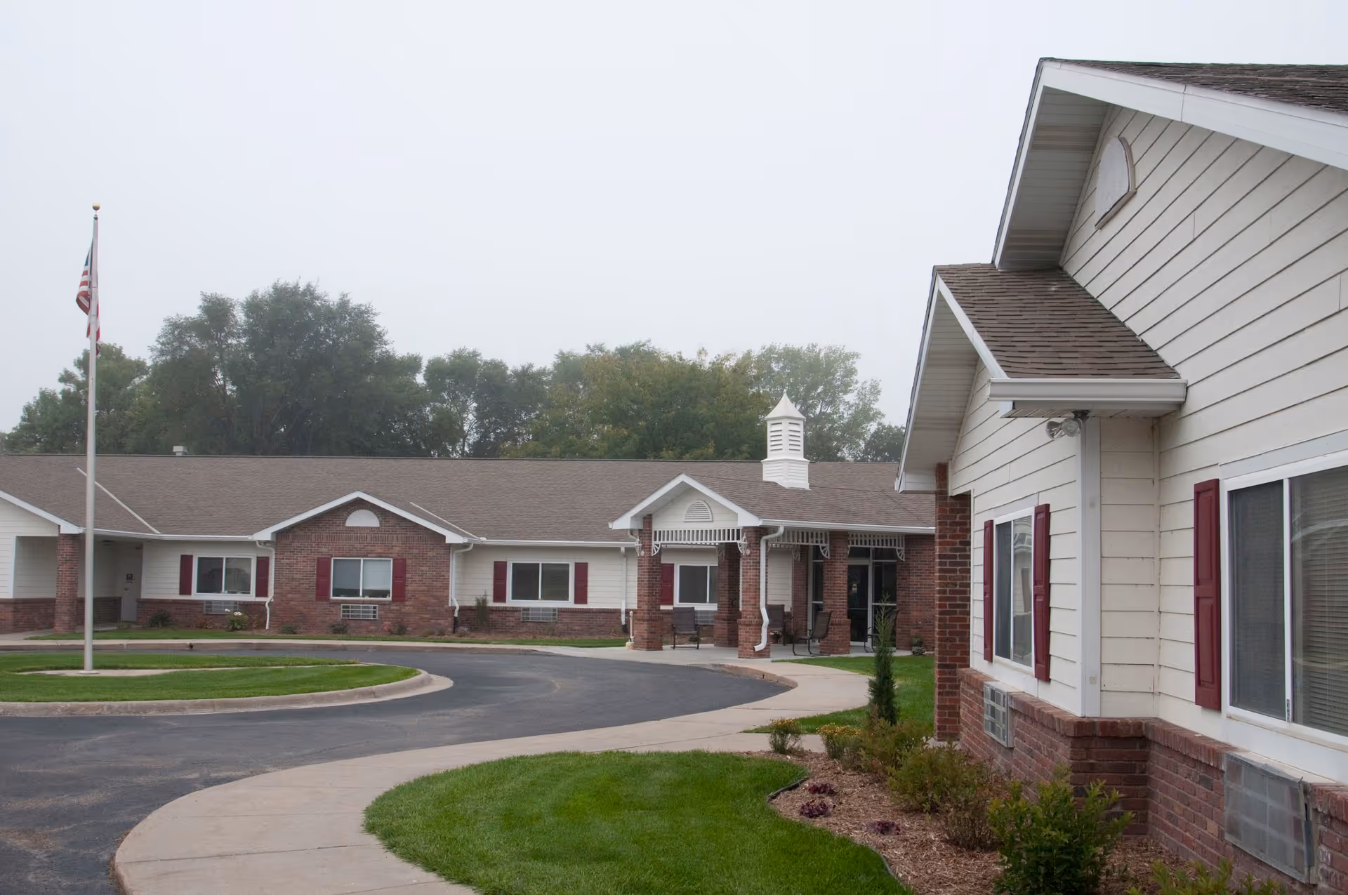 A single-story brick-and-siding assisted living building with a covered entrance, circular driveway, American flag, and landscaped lawn.