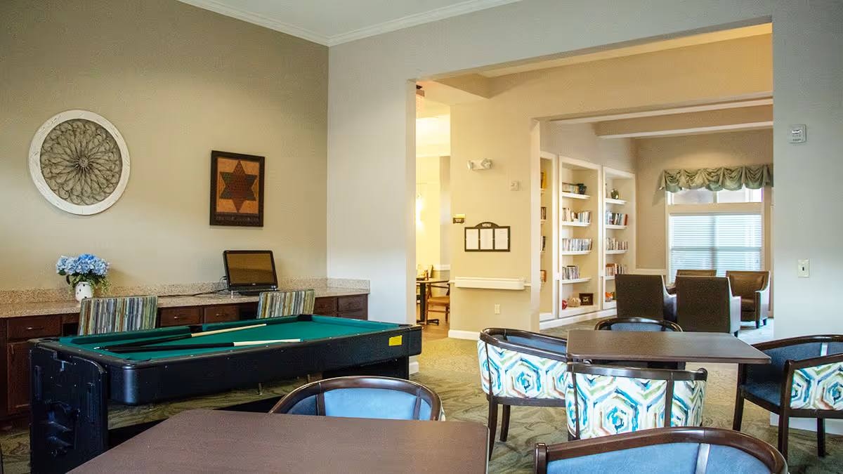 Interior view of a senior living facility common area featuring a pool table, tables with chairs, a computer on a counter, wall decorations, built-in bookshelves, and a seating area near a window with valance curtains.
