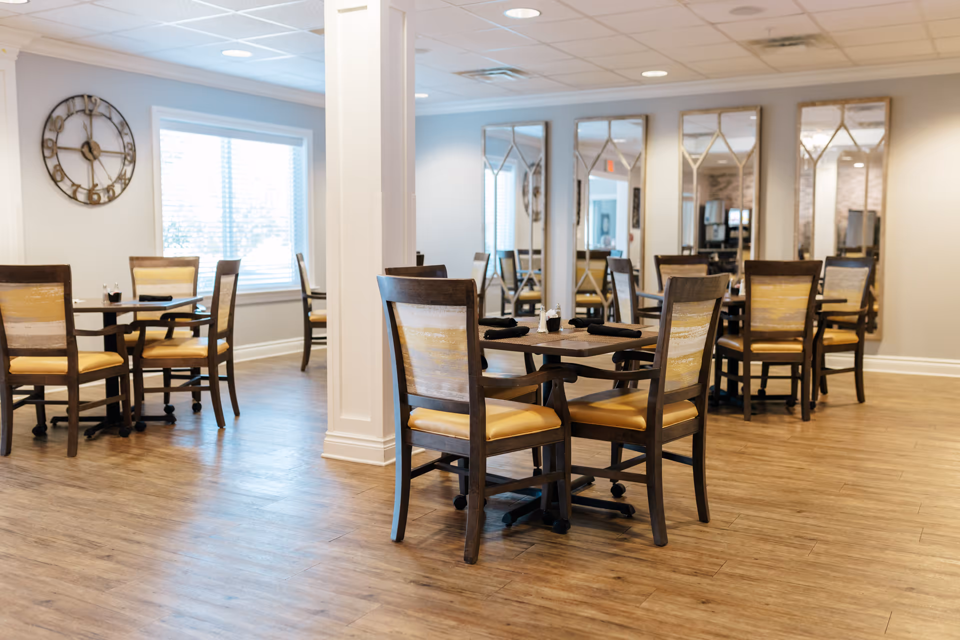 A bright dining room with wooden floors and several tables surrounded by wooden chairs with yellow cushions. The room features large windows with blinds, a decorative wall clock, and a series of tall mirrors on one wall reflecting the space.