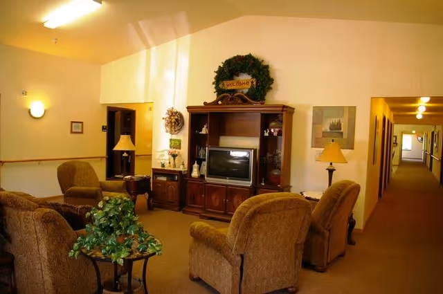 A cozy living room area in a senior living facility with four upholstered armchairs arranged around a small table with a green leafy plant. There is a wooden entertainment center with a television and decorative items, a wreath with a welcome sign above it, and two table lamps. A hallway with several doors and ceiling lights extends to the right.