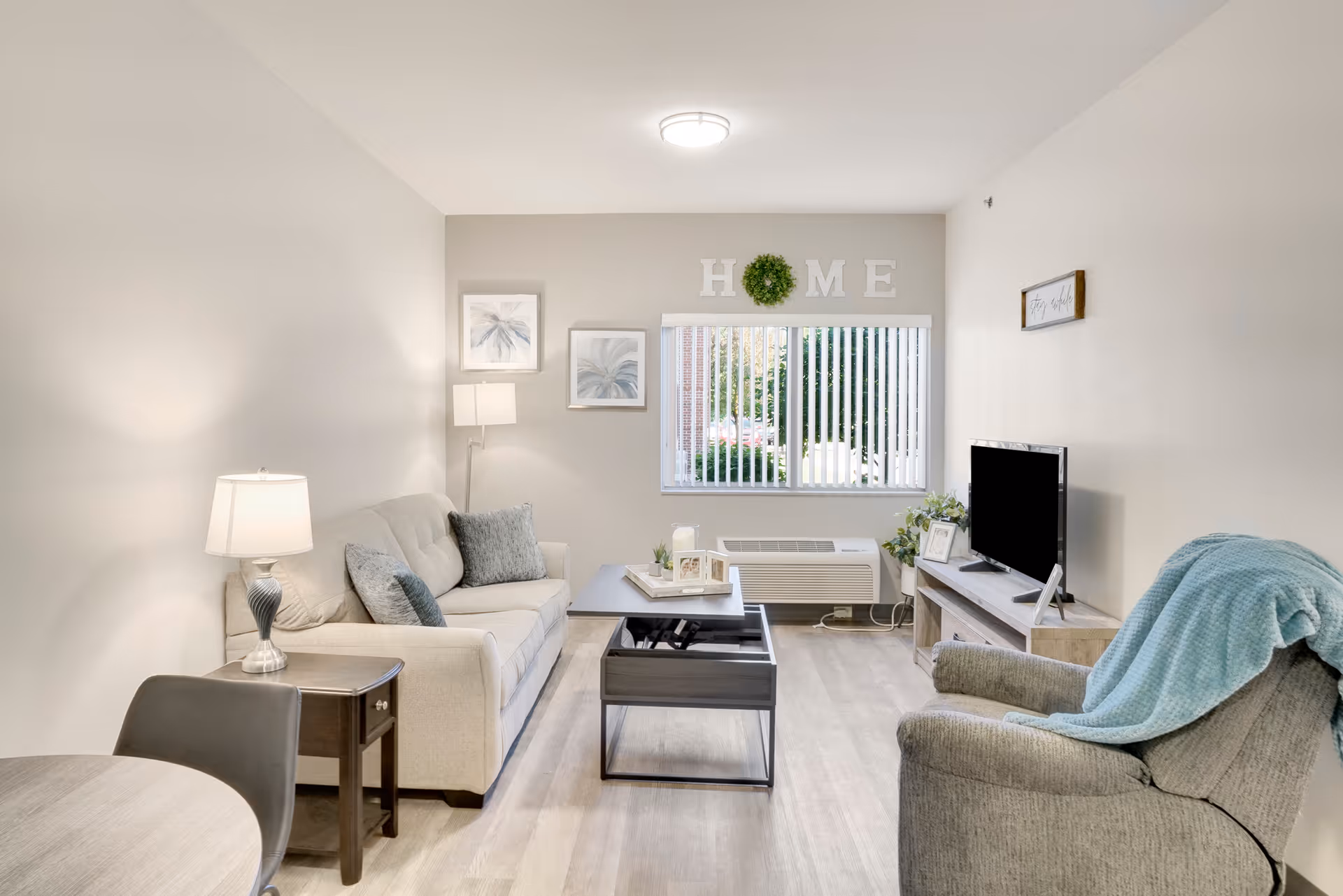 A bright and cozy living room with a beige sofa adorned with gray cushions, a gray armchair with a blue throw blanket, a wooden coffee table with a lift-top, and a TV on a wooden stand. The room has light-colored walls and flooring, a window with vertical blinds, and decorative wall art including the word 'HOME' with a green wreath in place of the letter 'O'. A table lamp and a floor lamp provide warm lighting.