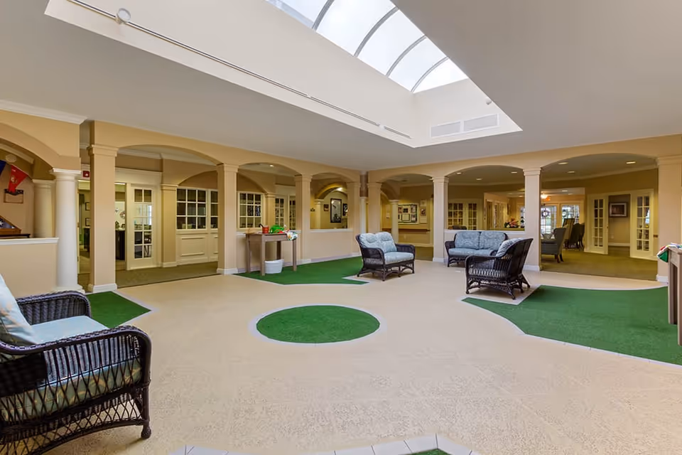 Indoor common area with beige walls and columns, featuring wicker chairs with cushions arranged around artificial green turf patches on the floor. The ceiling has a large skylight allowing natural light to brighten the space. Multiple doorways and windows lead to adjoining rooms.