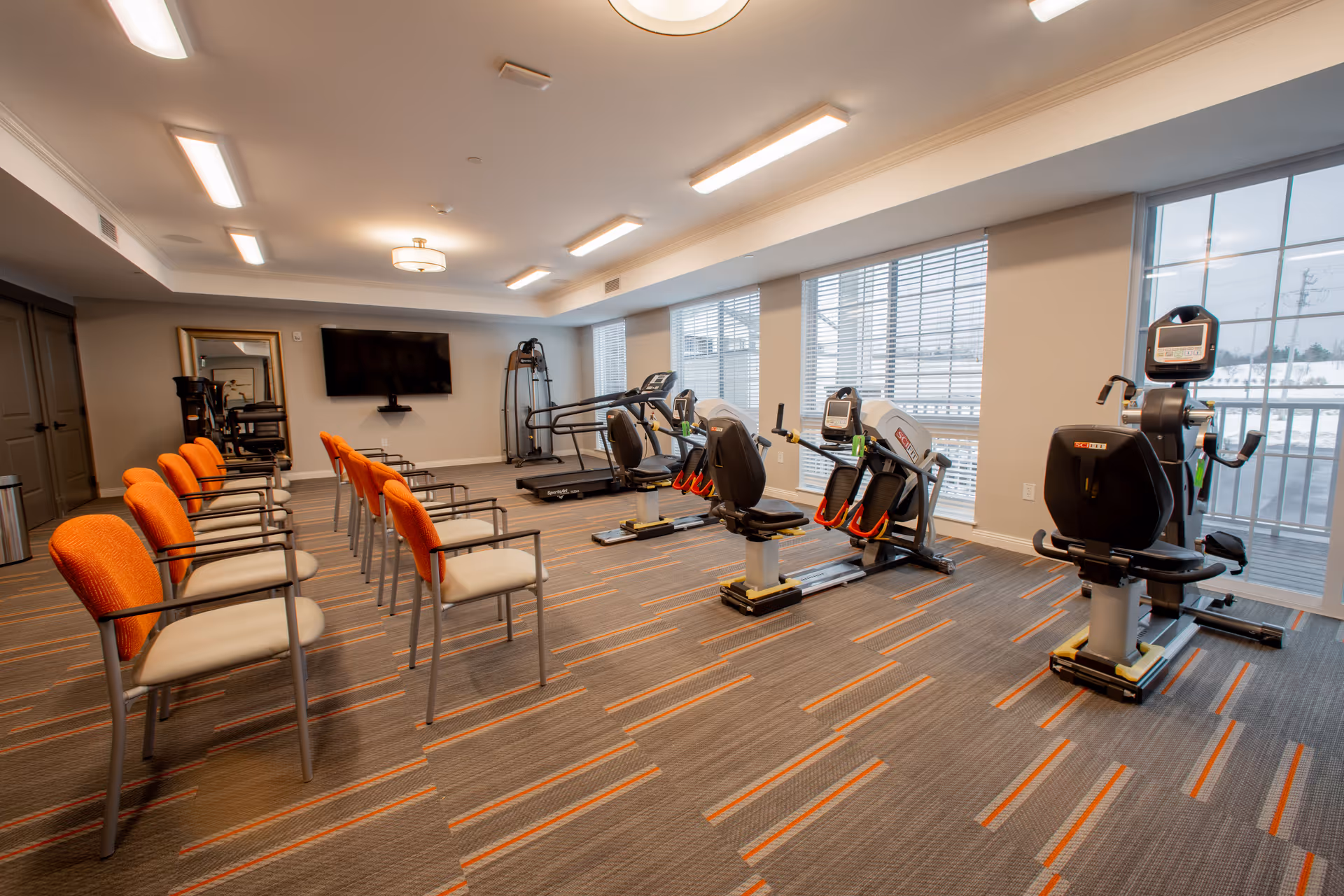Well-lit senior fitness room with exercise machines by the windows and a row of orange chairs.
