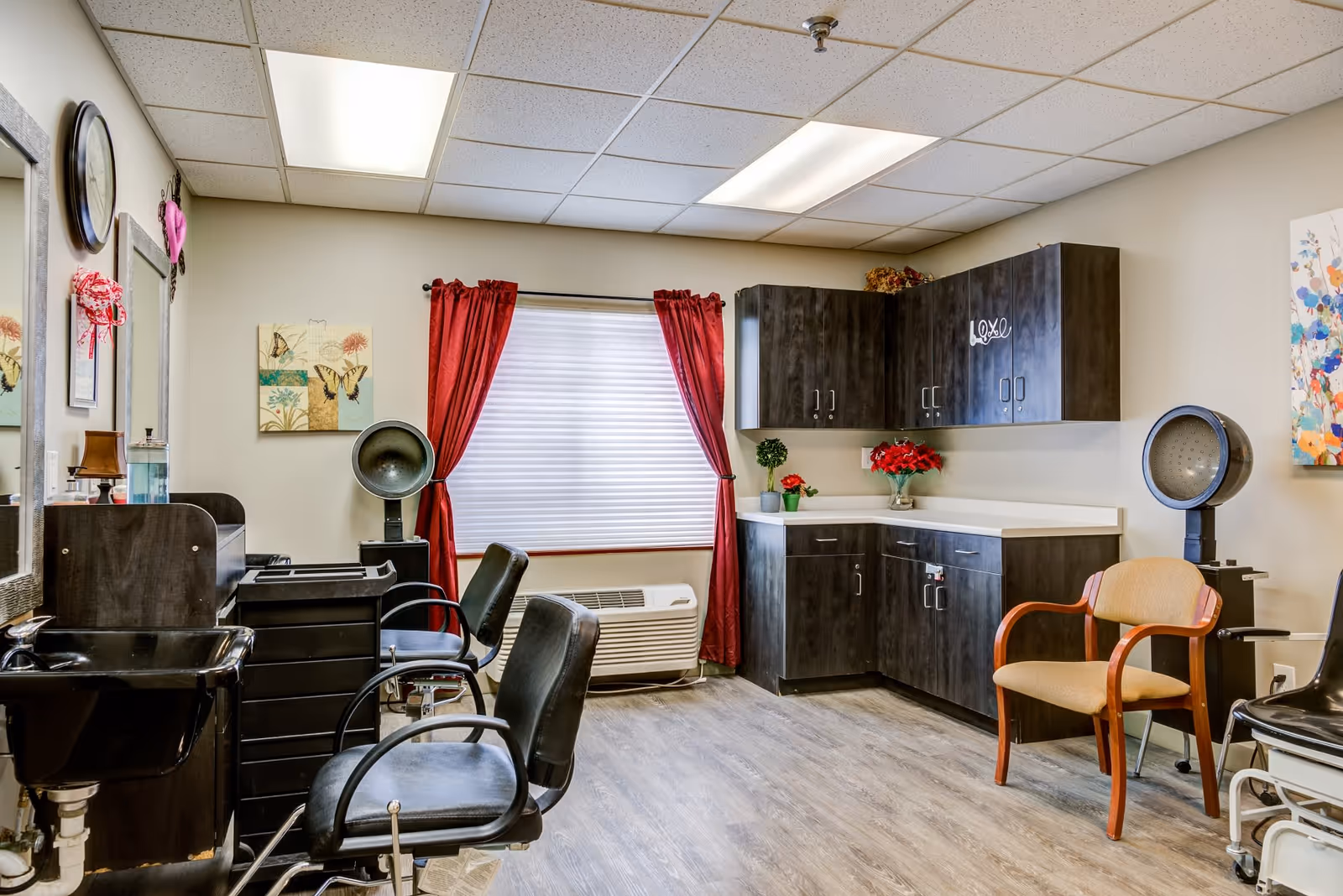 A bright salon-style room with styling chairs, hooded hair dryers, a sink, and cabinetry with a window and red curtains.