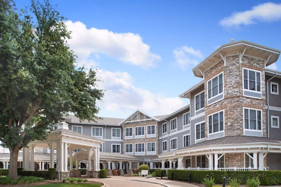 Exterior view of a multi-story senior living facility building with stone and gray siding, surrounded by landscaping including trees and bushes, under a blue sky with some clouds.
