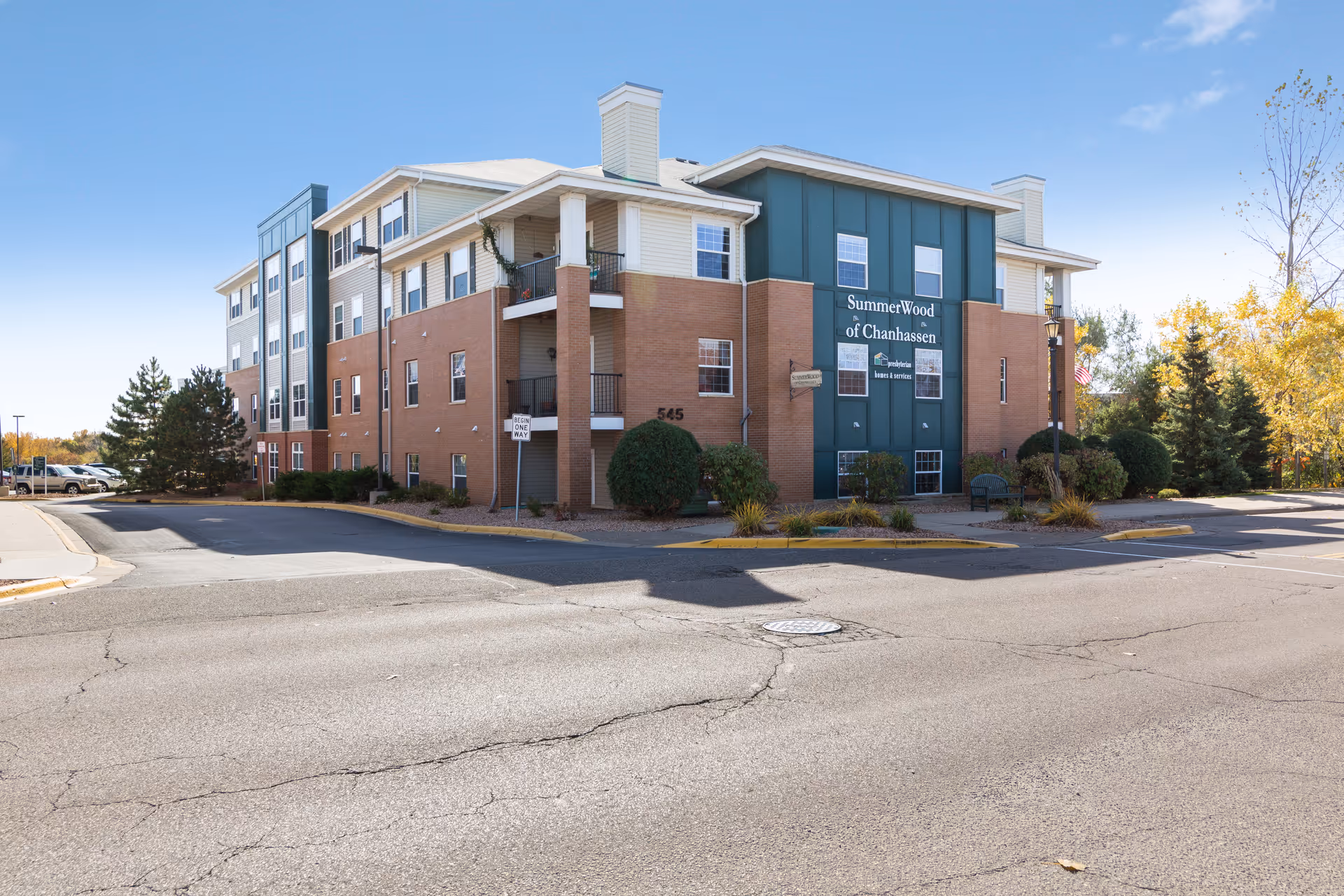 Exterior view of a multi-story senior living facility named SummerWood of Chanhassen, featuring brick and green paneling, with balconies and surrounding landscaping under a clear blue sky.