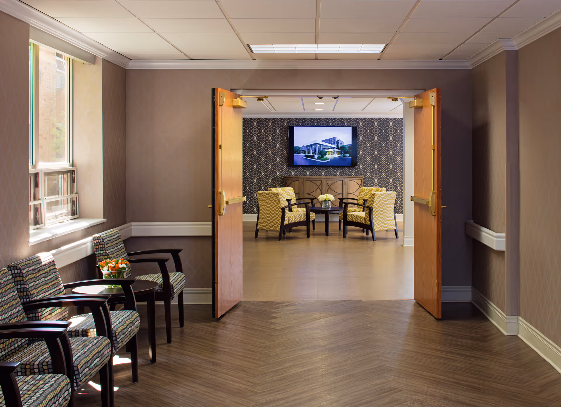 A hallway with patterned chairs and small tables with flowers on the left side, leading to an open double door entrance into a room with four yellow armchairs arranged around a small table with flowers. A flat screen TV is mounted on a patterned wall in the background.