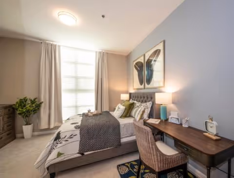 A cozy bedroom with a large window covered by light curtains, a bed with patterned bedding and multiple pillows, a wooden desk with a chair, two table lamps, and framed butterfly artwork on the wall.