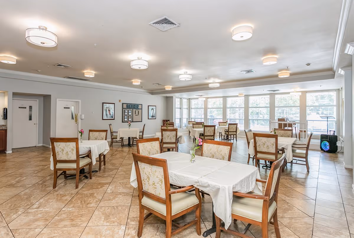 A spacious dining room with multiple tables covered in white tablecloths, each surrounded by wooden chairs with cushioned seats and floral upholstery. The room has large windows allowing natural light to fill the space, beige tiled floors, and several ceiling lights. There are framed pictures and a decorative sign on the far wall, and a speaker or sound system is visible near the windows.