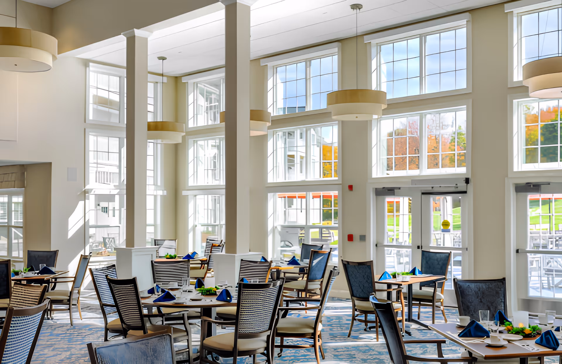 Bright and spacious dining room with large windows allowing natural light to fill the space. Multiple tables are set with blue napkins, plates, and glasses. Comfortable chairs surround the tables, and modern pendant lights hang from the ceiling. Outside the windows, greenery and a patio area are visible.