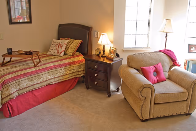 A cozy bedroom with a single bed covered in a striped red and beige bedspread, a wooden nightstand with a lamp, clock, and framed photo, and a beige armchair with a red pillow and throw blanket near a window with blinds.