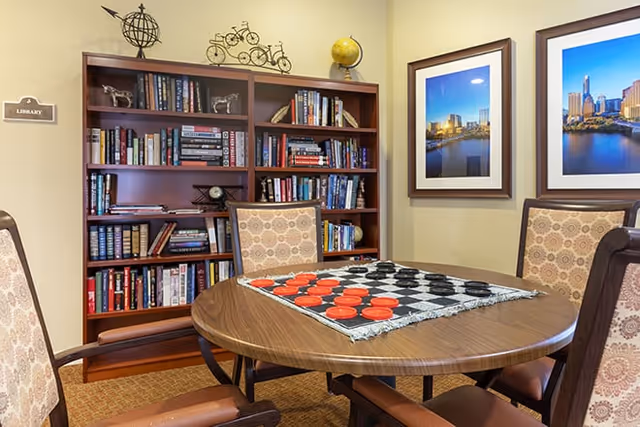 Round table with a checkers board and four upholstered chairs in a cozy room in front of a bookshelf labeled "Library" and framed city photos on the wall.