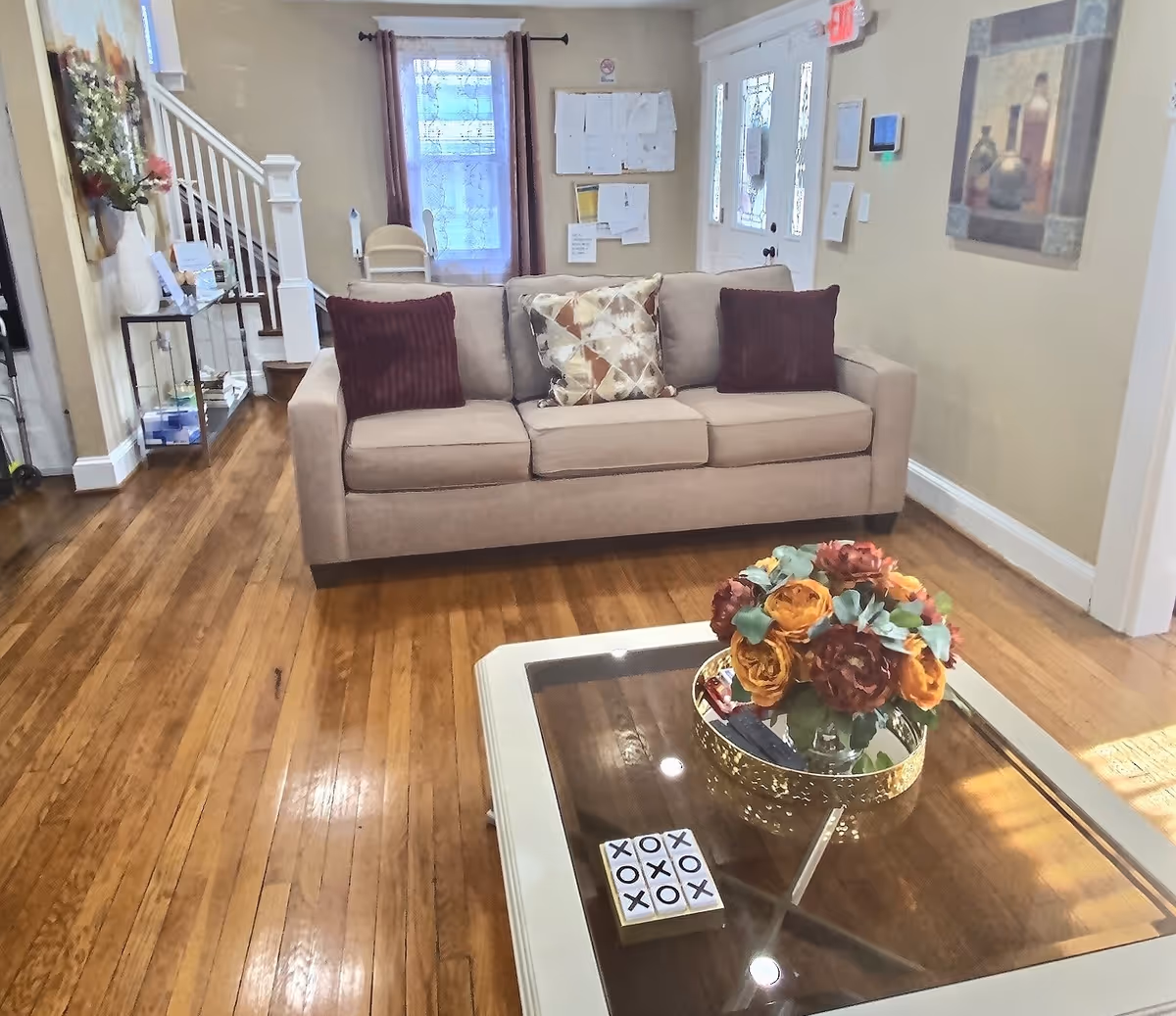 Bright living room with a beige sofa, decorative pillows, a glass-top coffee table with a floral arrangement, and hardwood floors leading to a front door and staircase.