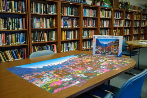A library room with wooden bookshelves filled with books along the walls. In the foreground, there is a wooden table with a partially completed colorful Ravensburger puzzle depicting a scenic mountain and flower landscape. Several blue chairs surround the table.