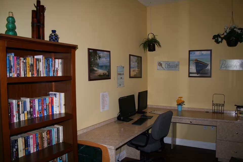 Small interior office area with a bookshelf, two computer monitors on a corner desk, framed beach artwork and hanging plants.