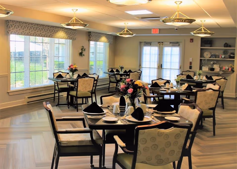 A bright dining room with several tables set for a meal, each table featuring black folded napkins, white plates, cups, and silverware. There are floral centerpieces on each table. The room has large windows with blinds allowing natural light to enter, light-colored walls, and modern ceiling lights. In the background, there is a double door with sheer curtains and a shelving unit with decorative items.