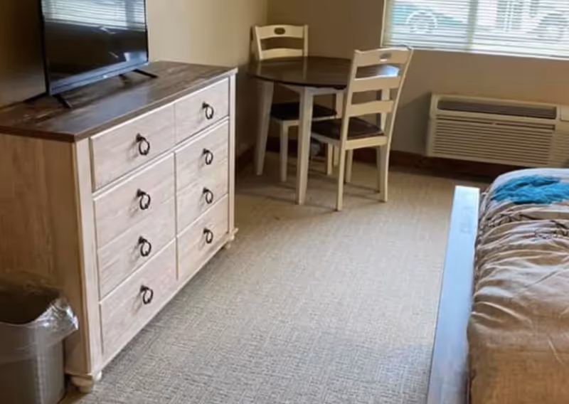 A room with a wooden dresser topped by a flat-screen TV, a small round table with two chairs near a window, and part of a bed with a brown comforter and a blue blanket. There is also a wall-mounted air conditioning unit below the window and a trash can beside the dresser.