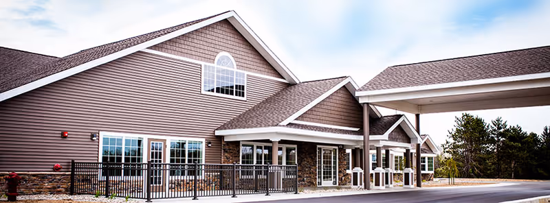 Exterior view of a single-story senior living facility building with brown siding, stone accents, multiple windows, and a covered entrance. There is a black metal fence enclosing a patio area and trees in the background under a partly cloudy sky.