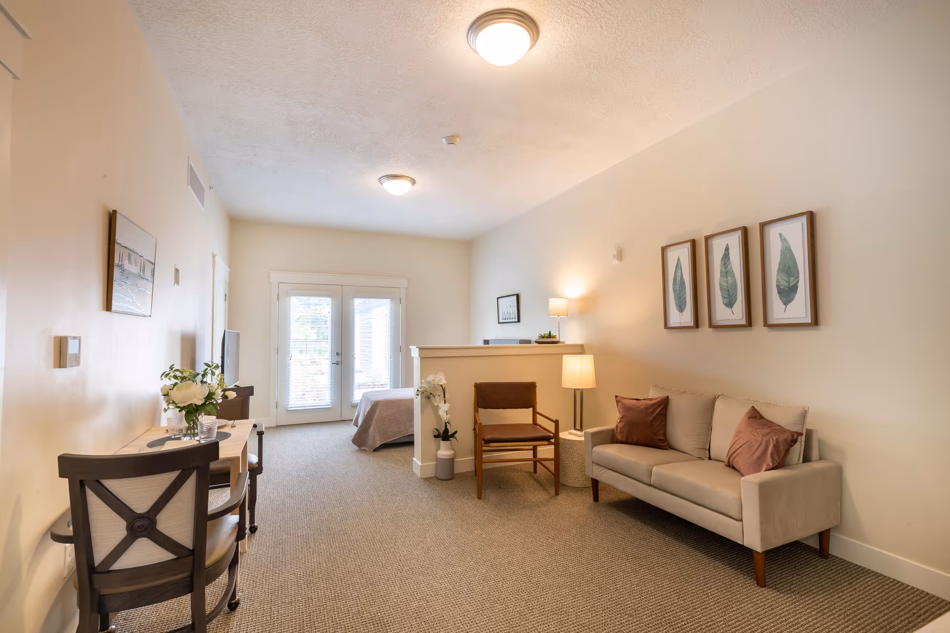 Bright studio-style interior with a sofa, chair, side table, small desk, and a bed near glass patio doors.