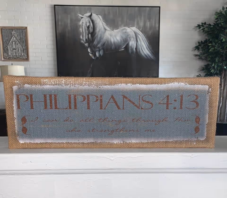 A decorative sign with the text 'PHILIPPIANS 4:13 I can do all things through Him who strengthens me' displayed on a shelf. Behind the sign, there is a black and white painting of a horse, a small framed artwork on a white brick wall, and a green leafy plant.