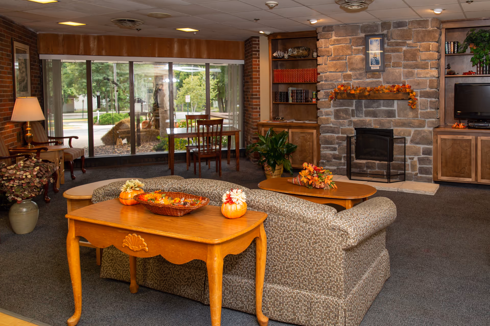 A cozy living room area with a patterned sofa facing a stone fireplace decorated with autumn leaves. There are wooden tables with fall-themed decorations, a lamp on a side table, a bookshelf with books and decor, a TV on a wooden cabinet, and large windows showing greenery outside.
