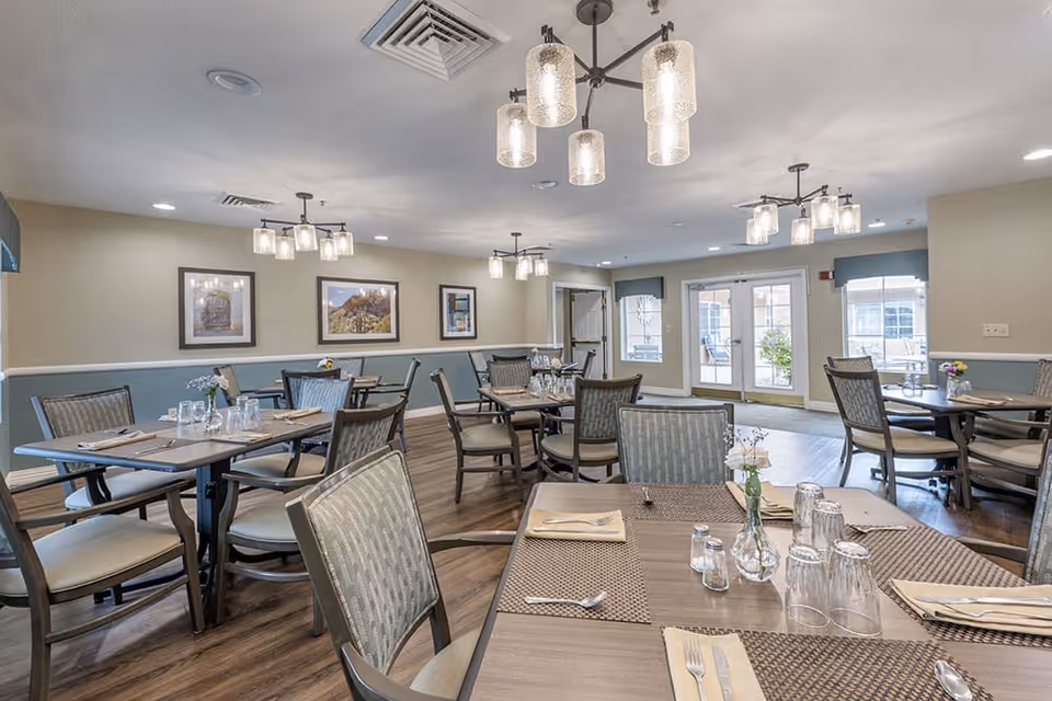 A dining room in a senior living facility with several tables set with placemats, napkins, silverware, glasses, and small flower vases. The room has wooden flooring, light-colored walls with blue wainscoting, framed artwork on the walls, and multiple ceiling light fixtures. Large windows and glass doors allow natural light to enter the space.