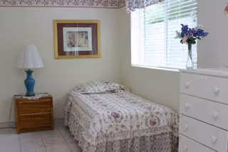 A small bedroom with a single bed covered in a floral bedspread, a wooden nightstand with a blue lamp, a framed picture on the wall, a window with blinds and a floral valance, and a white dresser with a vase of flowers on top.