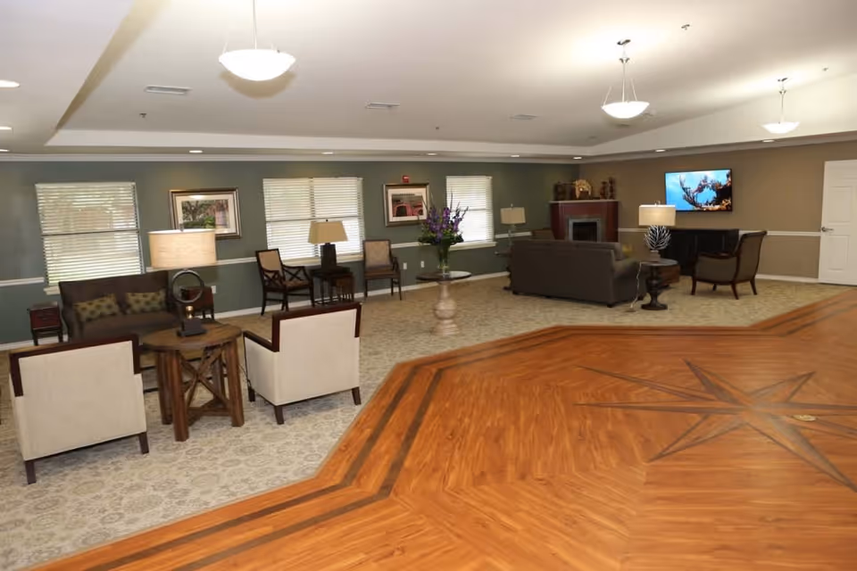 A spacious assisted living facility common area with a mix of carpet and wood flooring featuring a star design. The room includes multiple seating arrangements with sofas, armchairs, side tables, lamps, framed artwork on the walls, and a flat-screen TV mounted above a fireplace.