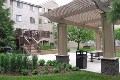 Outdoor patio area at Sterling Ponds featuring a wooden pergola, picnic tables, a trash can, landscaped greenery, and a multi-story residential building in the background.