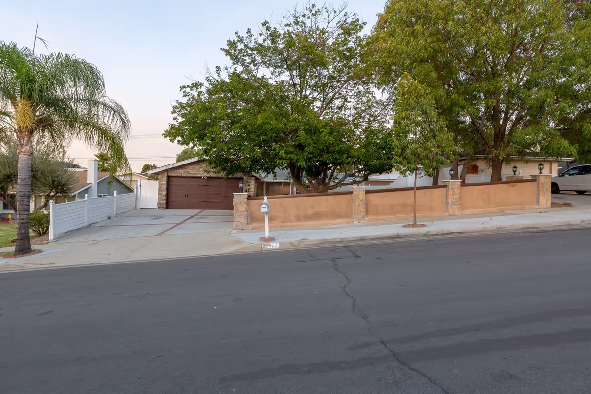 Street view of a single-story home with a two-car garage, driveway, low stucco wall and trees in front.