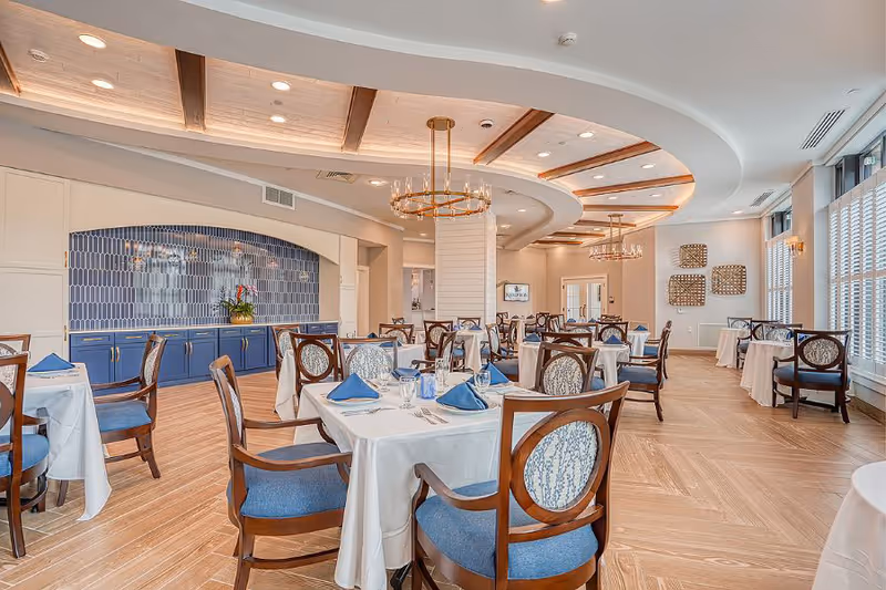 A bright and elegant dining room with multiple tables covered in white tablecloths, each set with blue napkins, glassware, and silverware. The room features wooden chairs with blue cushions and patterned backs, a wooden floor, large windows with white blinds, and modern chandeliers hanging from a ceiling with wooden accents. A blue tiled backsplash and cabinetry are visible along one wall.